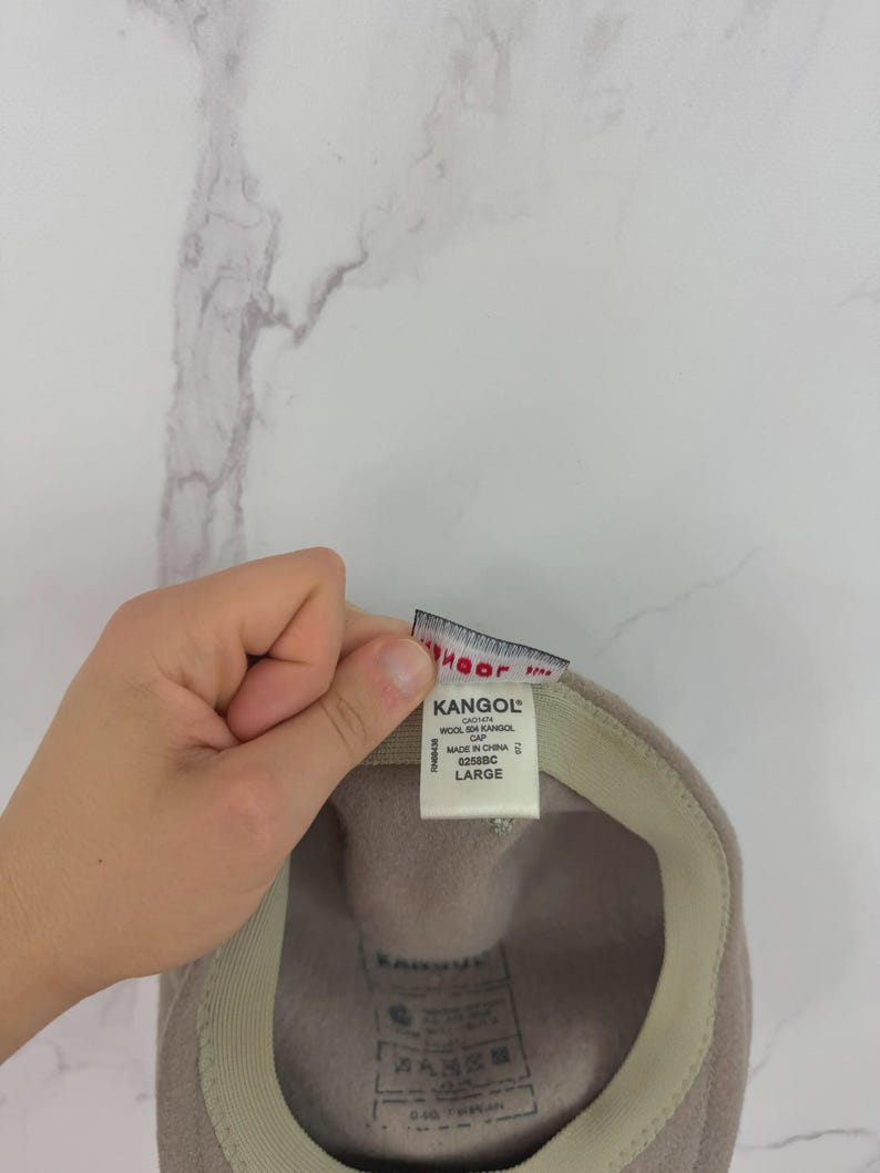 May include: A light brown Kangol wool cap with a beige interior. The cap has a tag that reads "Kangol" and "Large". The cap is made of wool and is a classic style.