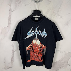 May include: Black t-shirt featuring the band name "Sodom" in blue and white stylized text. Below the text is a graphic of a skull with red and orange details. The shirt is hanging on a black hanger.