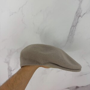 May include: A taupe-colored, flat cap made of a soft, textured material. The hat has a rounded crown and a small, stiff brim. The cap is held up by a person's arm. This classic style is suitable for various occasions.