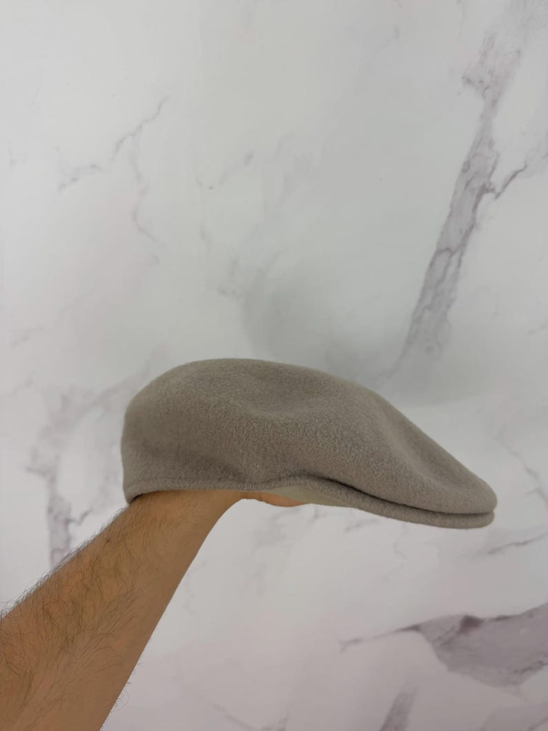 May include: A taupe-colored, flat cap made of a soft, textured material. The hat has a rounded crown and a small, stiff brim. The cap is held up by a person's arm. This classic style is suitable for various occasions.