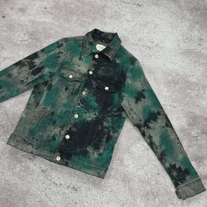 May include: A denim jacket with a tie-dye pattern in shades of green, black, and gray. The jacket features a classic collar, button-up front, two chest pockets, and buttoned cuffs. The garment is laid flat on a gray surface.