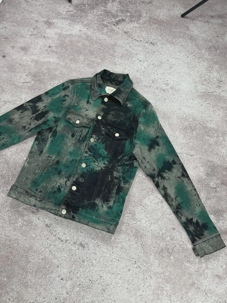 May include: A denim jacket with a tie-dye pattern in shades of green, black, and gray. The jacket features a classic collar, button-up front, two chest pockets, and buttoned cuffs. The garment is laid flat on a gray surface.