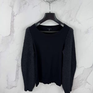 Könnte beinhalten: Schwarzer Pullover mit langen Ärmeln. Die Ärmel sind in einem dunkleren Grauton gehalten und haben eine flauschige Textur. Der Pullover hat einen runden Ausschnitt.