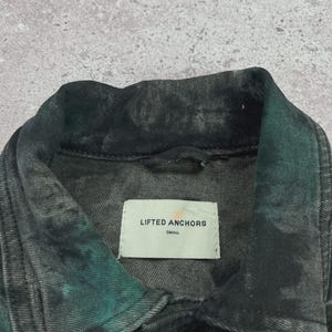 May include: Close-up of a denim jacket with a tie-dye effect in shades of black and teal. The jacket has a collar, a button closure, and a label that reads "LIFTED ANCHORS SMALL".