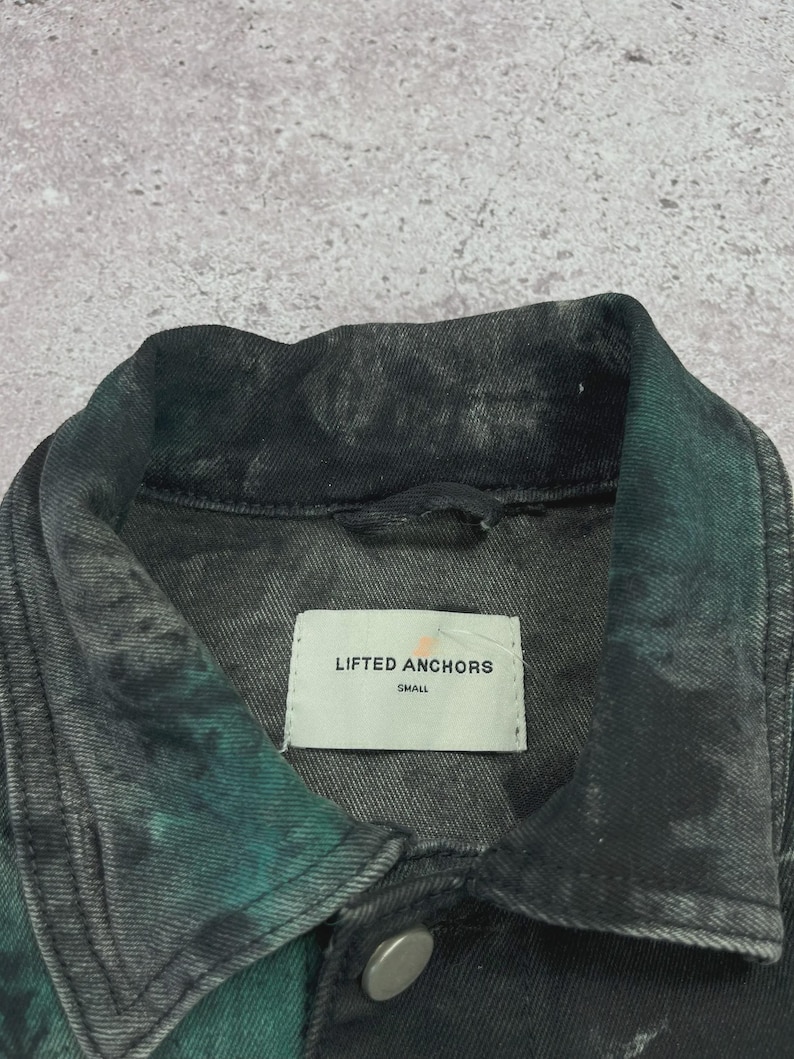 May include: Close-up of a denim jacket with a tie-dye effect in shades of black and teal. The jacket has a collar, a button closure, and a label that reads "LIFTED ANCHORS SMALL".