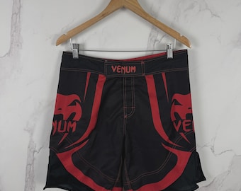 Könnte beinhalten: Schwarze und rote Sportshorts mit dem Venum-Logo und dem Markennamen in Rot auf beiden Beinen. Die Shorts haben einen breiten Bund mit einem zentralen Venum-Logo, rote Kontrastnähte und Seitenschlitze für Bewegungsfreiheit.