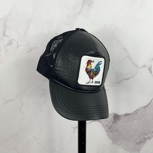 Puede incluir: Gorra de camionero de cuero negro con panel trasero de malla. La gorra presenta un parche de gallo colorido con la palabra "COQ" bordada debajo.