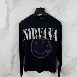 May include: Black sweater with the word "NIRVANA" in white block letters across the chest. Below the text is a purple smiley face with crossed-out eyes and a protruding tongue. The sweater has long sleeves and a crew neck.
