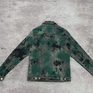May include: A denim jacket with a tie-dye pattern in shades of green and black. The jacket has a classic design with a collar, button closures, and pockets. The back of the jacket is visible, showcasing the unique dye pattern.