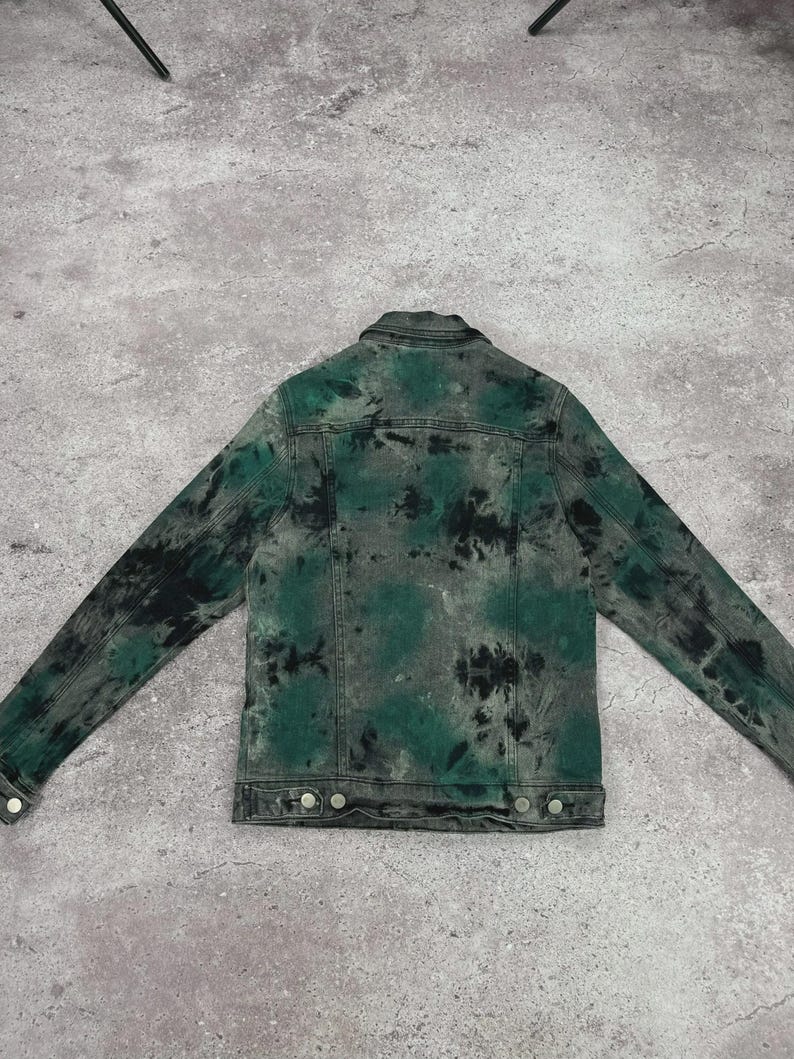 May include: A denim jacket with a tie-dye pattern in shades of green and black. The jacket has a classic design with a collar, button closures, and pockets. The back of the jacket is visible, showcasing the unique dye pattern.