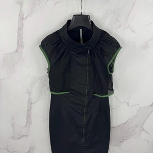 May include: Black sleeveless dress with a high collar and a full-length zip. The dress has a mesh overlay with green trim around the shoulders. It hangs on a black hanger against a marble background.