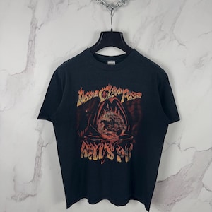 Peut inclure: T-shirt noir avec un motif graphique avec le texte "Insane Clown Posse" et "Hell's Pit" dans un style gothique enflammé. Le motif comprend une figure encapuchonnée et des flammes. Le t-shirt est suspendu à un cintre noir.