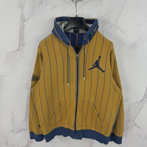 May include: Mustard yellow zip-up hoodie with navy blue pinstripes and accents. Features a front pocket and the Jumpman logo on the chest. The hood and cuffs are navy blue. A classic athletic-inspired design.