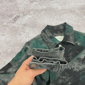 May include: A denim jacket with a unique tie-dye effect in shades of green and gray. The jacket features a classic collar, buttoned cuffs, and a button closure. The fabric has a textured appearance.