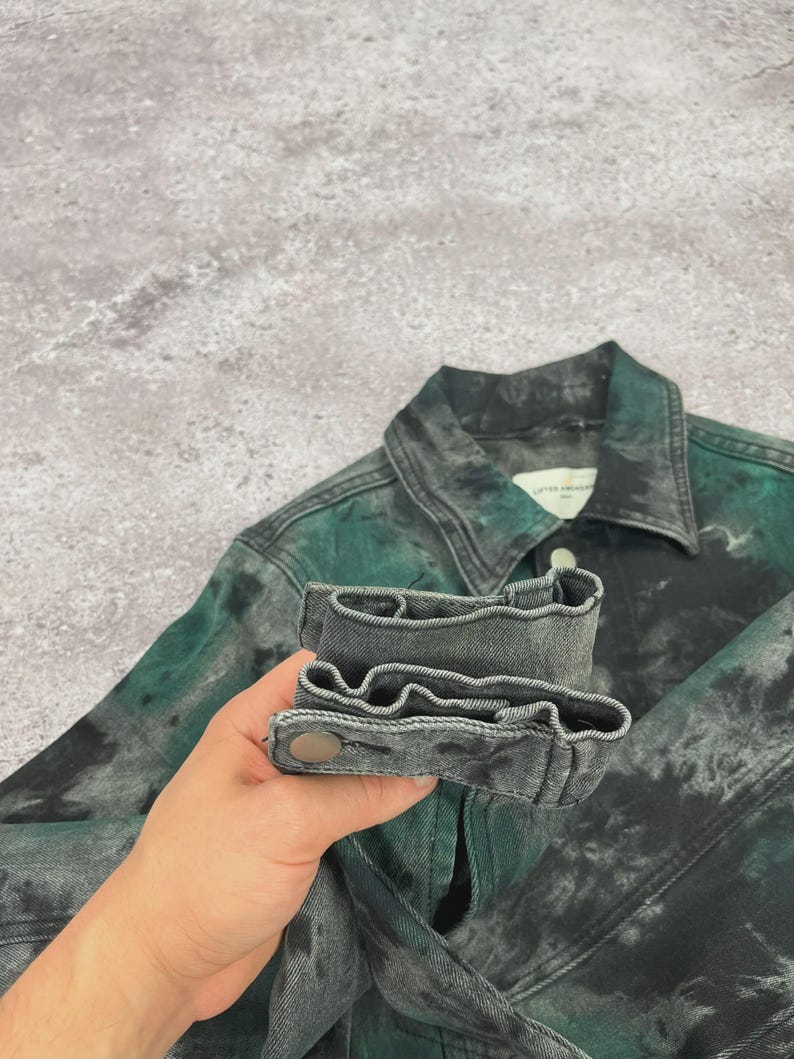 May include: A denim jacket with a unique tie-dye effect in shades of green and gray. The jacket features a classic collar, buttoned cuffs, and a button closure. The fabric has a textured appearance.