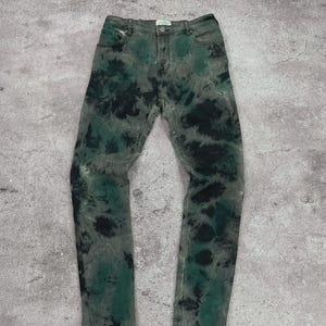 May include: Green and black tie-dye jeans. The jeans have a classic five-pocket design with a button and zipper closure. The tie-dye pattern is a mix of dark green and black on a gray denim base.