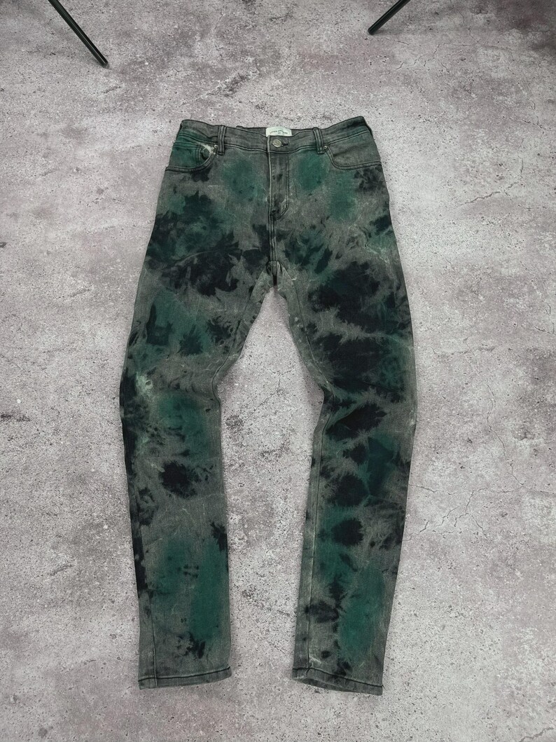 May include: Green and black tie-dye jeans. The jeans have a classic five-pocket design with a button and zipper closure. The tie-dye pattern is a mix of dark green and black on a gray denim base.