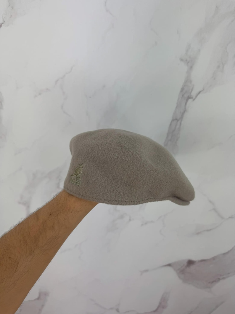 May include: A light gray Kangol beret with a textured, fuzzy surface. The beret features the Kangol logo embroidered in a darker gray on the side. This classic hat is a stylish accessory for any outfit.