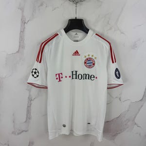 May include: White Adidas soccer jersey with red stripes on the shoulders and sleeves. The jersey features the Bayern Munich team logo, the Adidas logo, and the T-Home sponsor logo. A soccer ball graphic is on the left sleeve.