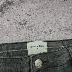 May include: Close-up of a pair of dark gray denim jeans. The jeans have a button closure and a label that reads "LIFTED ANCHORS 40". The fabric has a textured appearance.