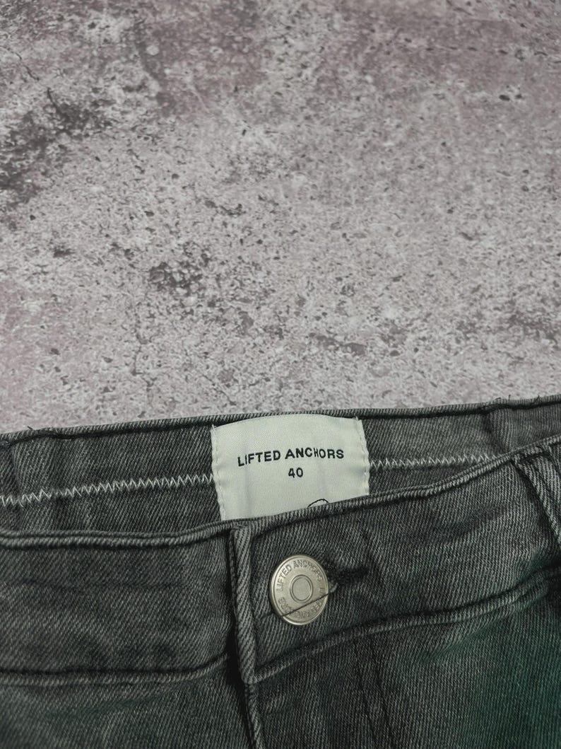 May include: Close-up of a pair of dark gray denim jeans. The jeans have a button closure and a label that reads "LIFTED ANCHORS 40". The fabric has a textured appearance.