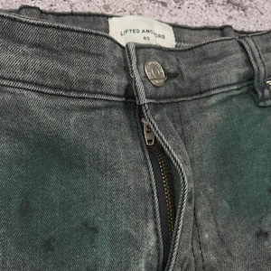 May include: Close-up of gray denim jeans with a zipper and button closure. The jeans have a faded, distressed look with dark spots. The brand "LIFTED ANCHORS" is visible on the label.
