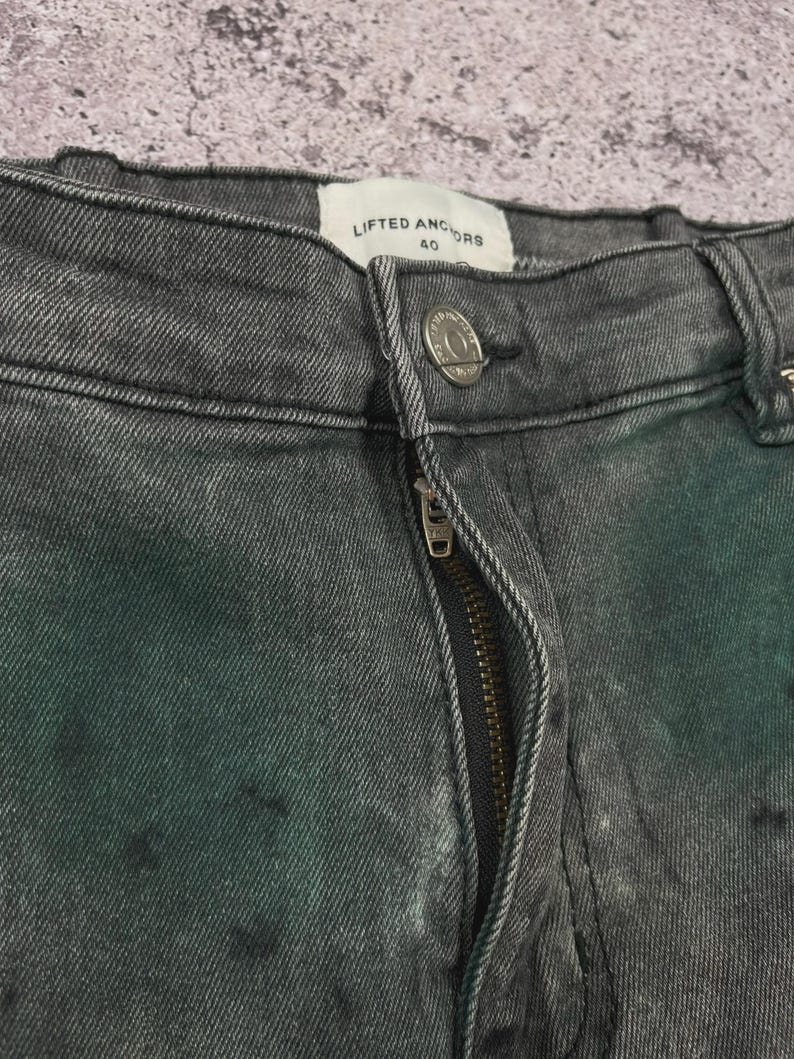 May include: Close-up of gray denim jeans with a zipper and button closure. The jeans have a faded, distressed look with dark spots. The brand "LIFTED ANCHORS" is visible on the label.