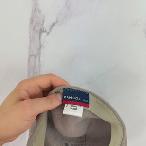 May include: A beige Kangol beret hat with a blue and red tag that reads "KANGOL blue" and "LARGE". The hat has a beige interior band and a soft, felt-like texture. The hat is a size large.