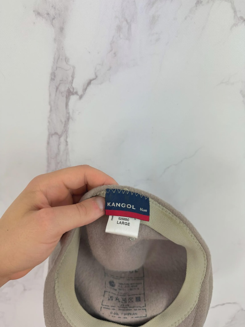 May include: A beige Kangol beret hat with a blue and red tag that reads "KANGOL blue" and "LARGE". The hat has a beige interior band and a soft, felt-like texture. The hat is a size large.