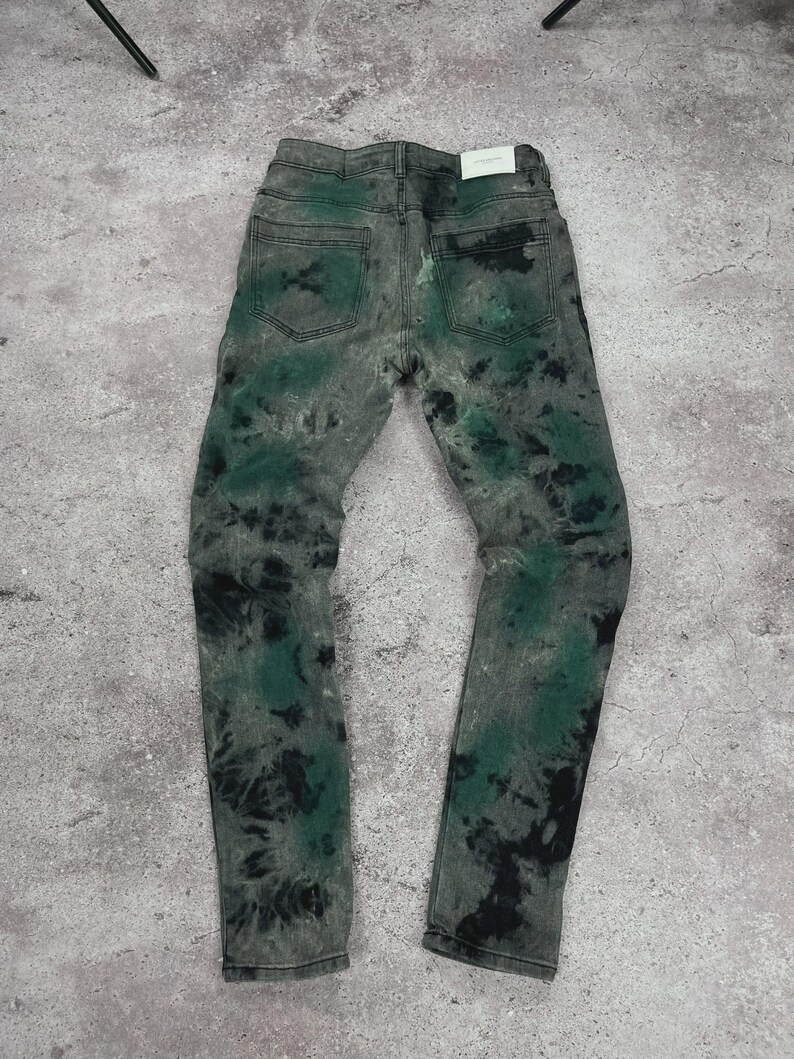 May include: Gray denim jeans with a tie-dye pattern in black and green. The jeans have a classic five-pocket design and a straight leg fit. The back pockets are visible, and a white tag is at the back.