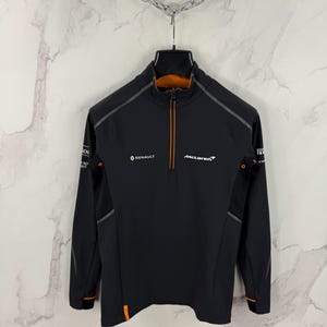 Può includere: Giacca nera con zip e dettagli arancioni e grigi. Presenta i loghi Renault e McLaren. La giacca ha un collo alto e una zip frontale. Le maniche hanno strisce grigie e dettagli arancioni.