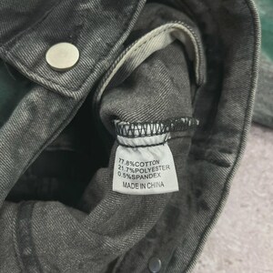 May include: Close-up of a gray denim jacket with a faded, distressed appearance. The jacket has a silver button and a tag with fabric composition details: 77.8% cotton, 21.7% polyester, and 0.5% spandex. The tag also indicates it was made in China.