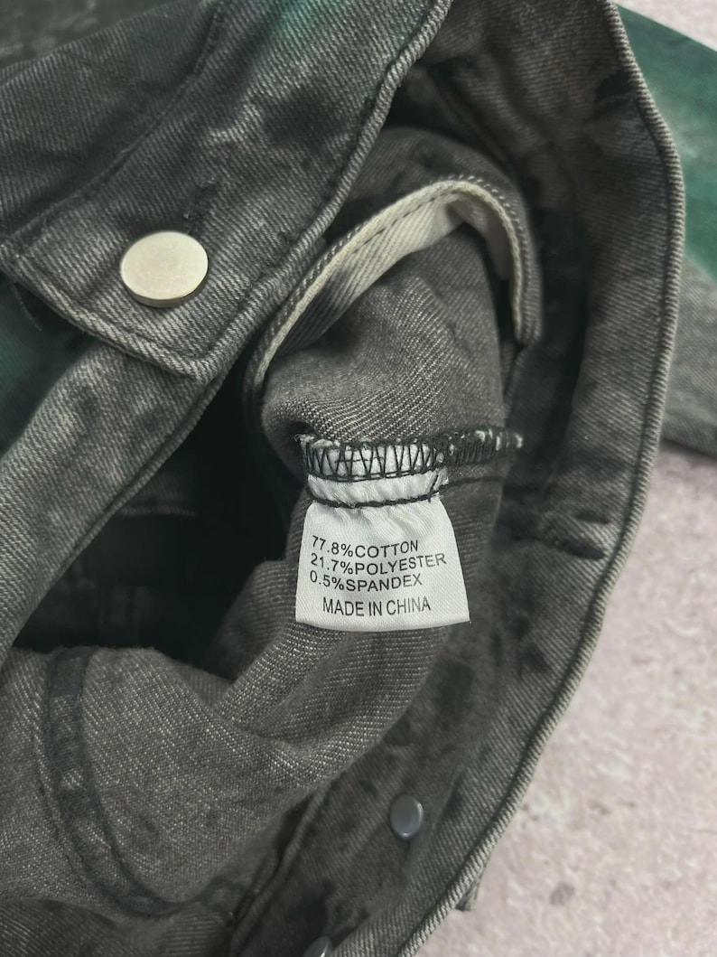 May include: Close-up of a gray denim jacket with a faded, distressed appearance. The jacket has a silver button and a tag with fabric composition details: 77.8% cotton, 21.7% polyester, and 0.5% spandex. The tag also indicates it was made in China.