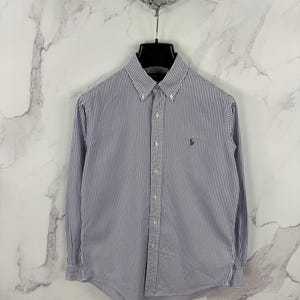 Puede incluir: Camisa abotonada a rayas verticales azules y blancas. La camisa de manga larga tiene cuello abotonado y un pequeño logotipo bordado en el pecho. La camisa está colgada de una percha negra.