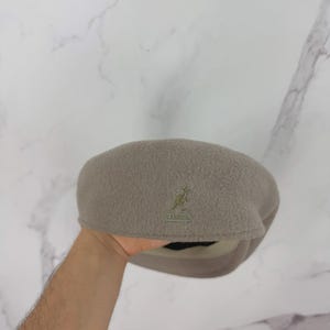 May include: A taupe-colored Kangol beret hat. The hat features a small embroidered kangaroo logo on the front. The beret is made of a soft, textured material and has a rounded crown and a fitted brim. The hat is a neutral color.