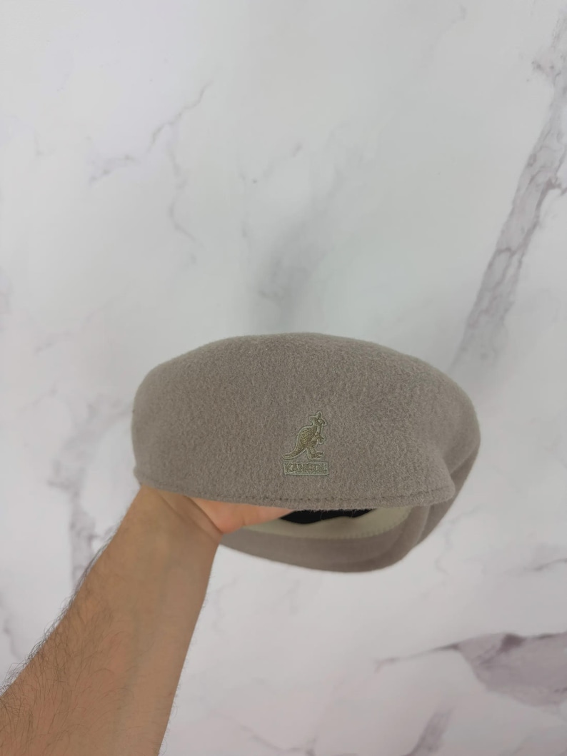 May include: A taupe-colored Kangol beret hat. The hat features a small embroidered kangaroo logo on the front. The beret is made of a soft, textured material and has a rounded crown and a fitted brim. The hat is a neutral color.