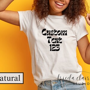 May include: A white t-shirt with the words "Custom Text 123" in black, displayed in a bold, retro font. The shirt is worn by a person with curly hair, against a yellow background. The word "Natural" is in a black box.