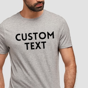 May include: A light gray t-shirt with the words "CUSTOM TEXT" printed in bold black letters. The shirt is short-sleeved and appears to be made of a soft, comfortable material. The image shows the upper torso of a person wearing the shirt.