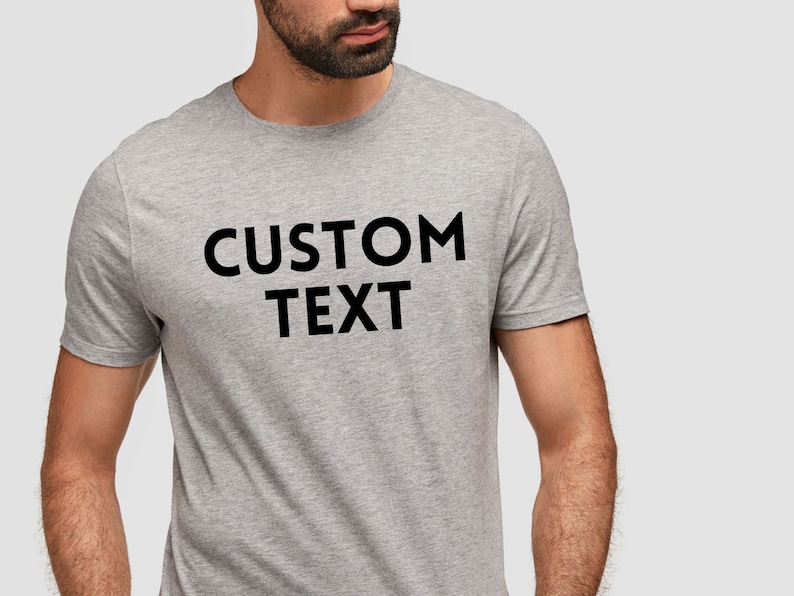May include: A light gray t-shirt with the words "CUSTOM TEXT" printed in bold black letters. The shirt is short-sleeved and appears to be made of a soft, comfortable material. The image shows the upper torso of a person wearing the shirt.