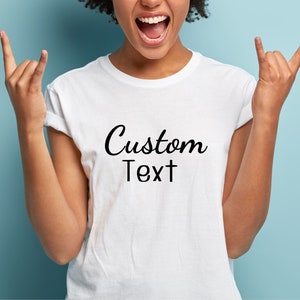 May include: White t-shirt with the words "Custom Text" in a black script font. The shirt is being worn by a person with their arms raised in a rock and roll gesture. The background is a solid light blue.
