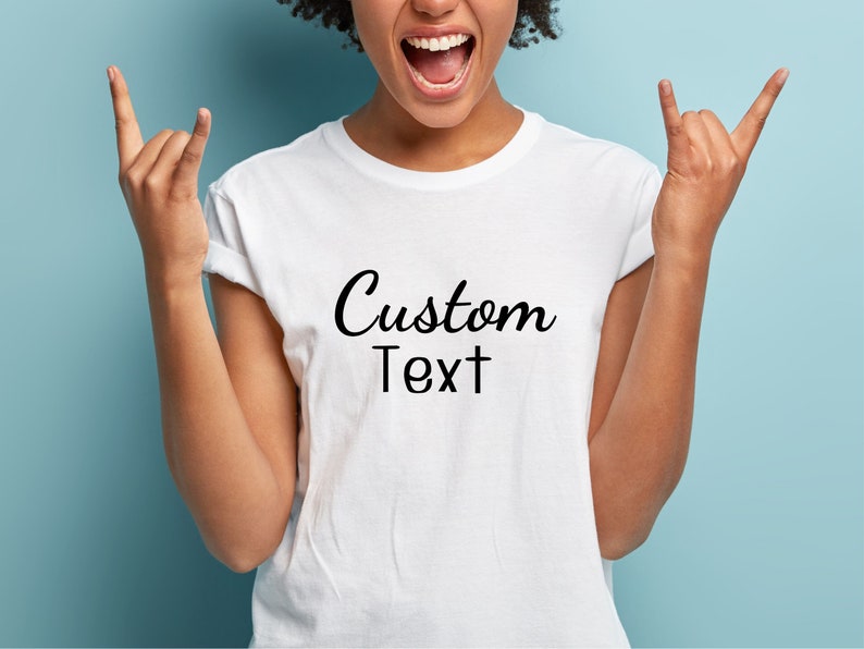 May include: White t-shirt with the words "Custom Text" in a black script font. The shirt is being worn by a person with their arms raised in a rock and roll gesture. The background is a solid light blue.