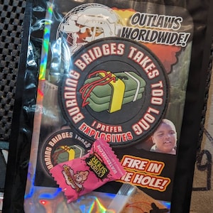 Puede incluir: Una bolsa negra con un interior holográfico iridiscente. La bolsa contiene un parche redondo negro con el texto "Burning Bridges Takes Too Long I Prefer Explosives". También hay un caramelo rosa con el texto "Extreme Sour Watermelon", una pequeña bolsa transparente con polvo rojo y una pegatina con el texto "Outlaws Worldwide! Fire in the Hole!"