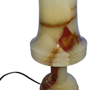 May include: A decorative table lamp made of onyx stone. The lamp features a cream-colored shade with red and brown veining, a flared base, and a round foot. A black power cord extends from the base.