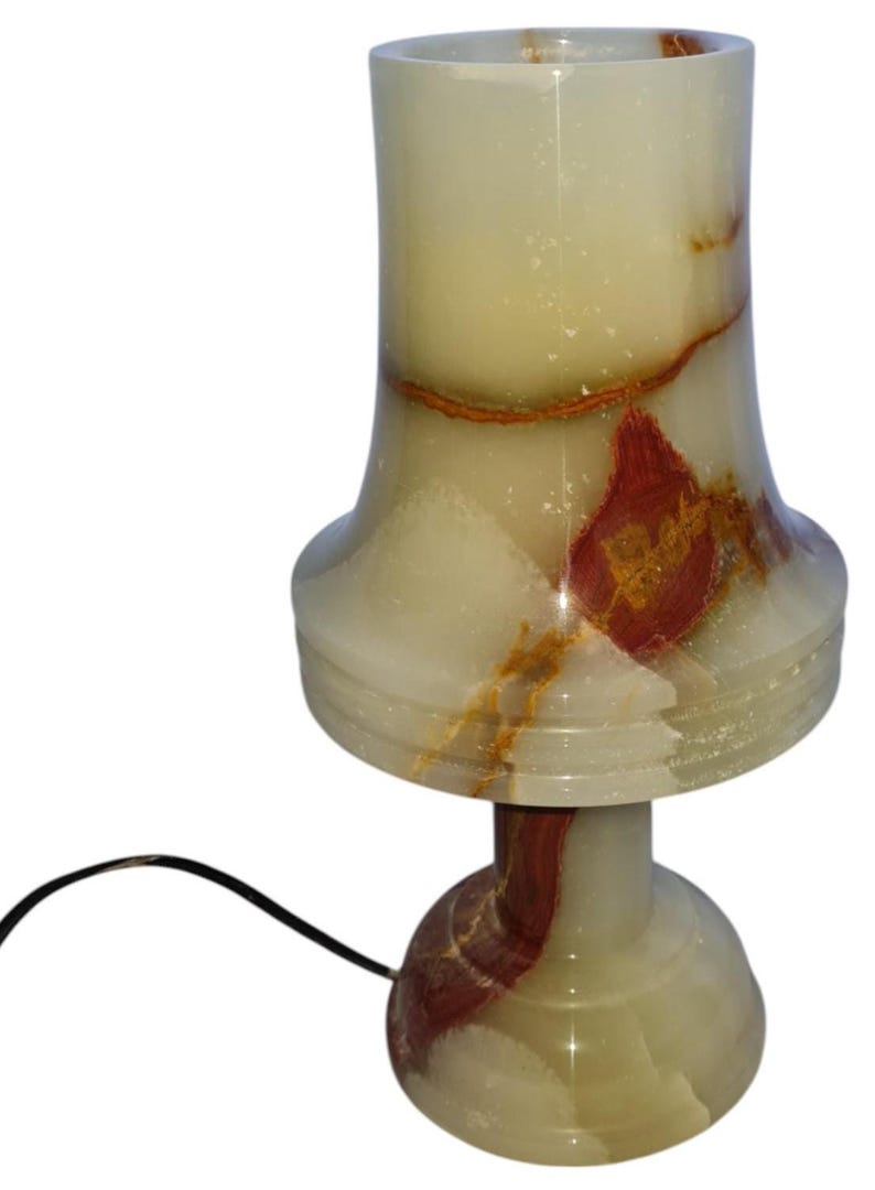 May include: A decorative table lamp made of onyx stone. The lamp features a cream-colored shade with red and brown veining, a flared base, and a round foot. A black power cord extends from the base.