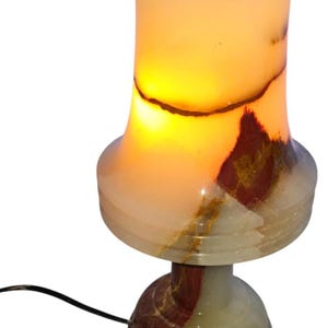 May include: A glowing onyx table lamp with a cylindrical shade and a rounded base. The lamp features a warm, amber glow emanating from within, with natural veining in shades of brown, red, and white. The lamp is connected to a black power cord.