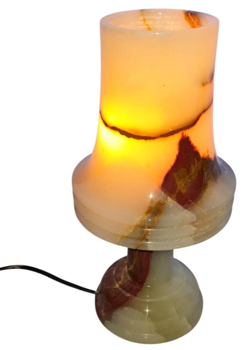 May include: A glowing onyx table lamp with a cylindrical shade and a rounded base. The lamp features a warm, amber glow emanating from within, with natural veining in shades of brown, red, and white. The lamp is connected to a black power cord.