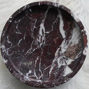 May include: A round, marble tray with a deep burgundy base and white veining. The tray has a smooth, polished surface and a slightly raised edge. The natural stone pattern creates a unique, decorative piece.