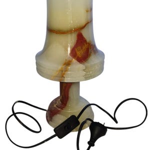 May include: A decorative table lamp made of translucent onyx stone. The lamp features a cream-colored shade with red and brown veining, a pedestal base, and a black power cord with an on/off switch. The lamp is approximately 10 inches tall.