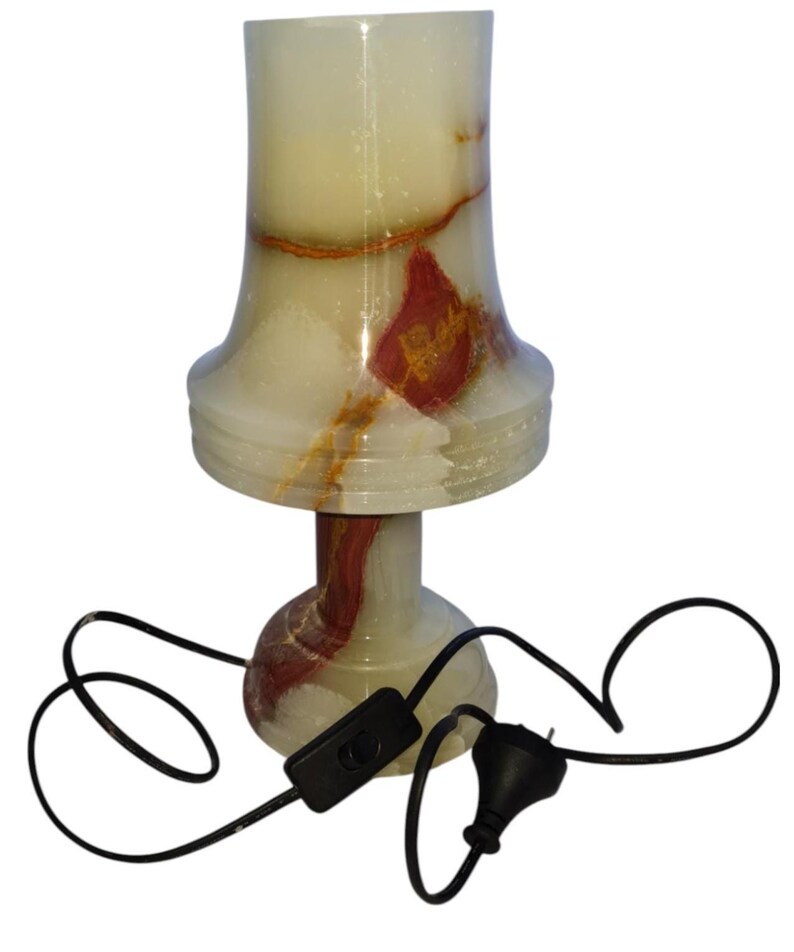 May include: A decorative table lamp made of translucent onyx stone. The lamp features a cream-colored shade with red and brown veining, a pedestal base, and a black power cord with an on/off switch. The lamp is approximately 10 inches tall.