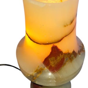 May include: A lit onyx lamp with a warm glow. The lamp has a bulbous base and a cylindrical top, with natural brown and red veining. The lamp is plugged in and ready to use.